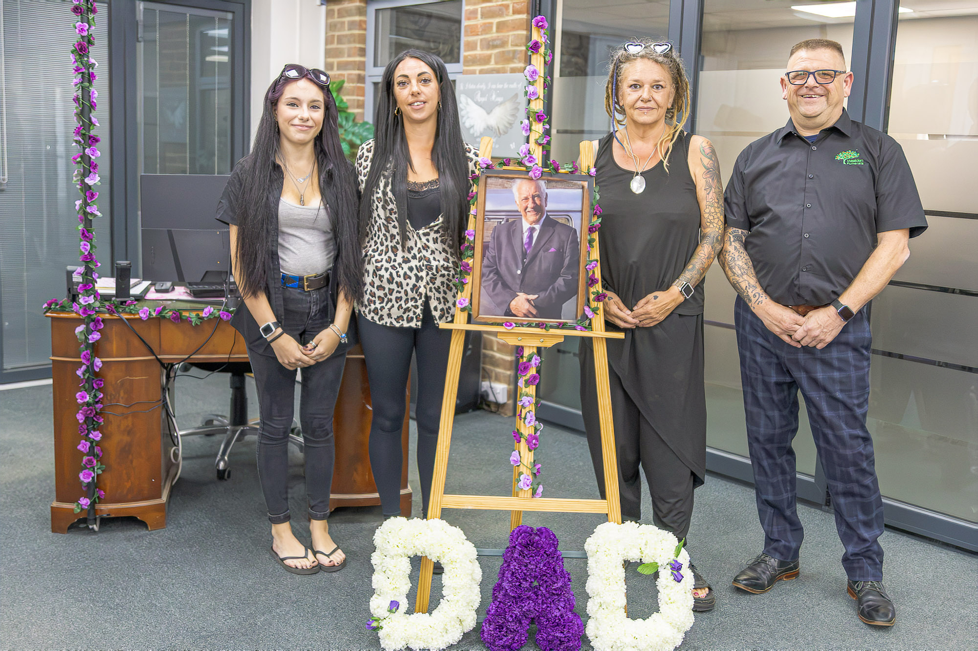A family with Andy beside a personal memorial portrait and floral tributes